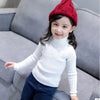 A young girl wearing a white knit turtleneck sweater and a red knitted hat sitting on a gray sofa.