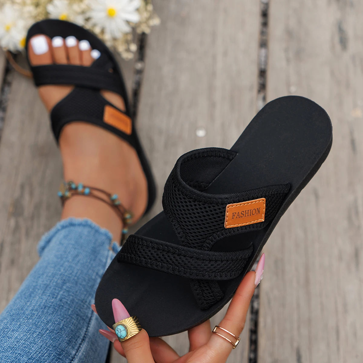 A hand holds a black breathable mesh flat sandal with a cushioned sole against a wooden background.