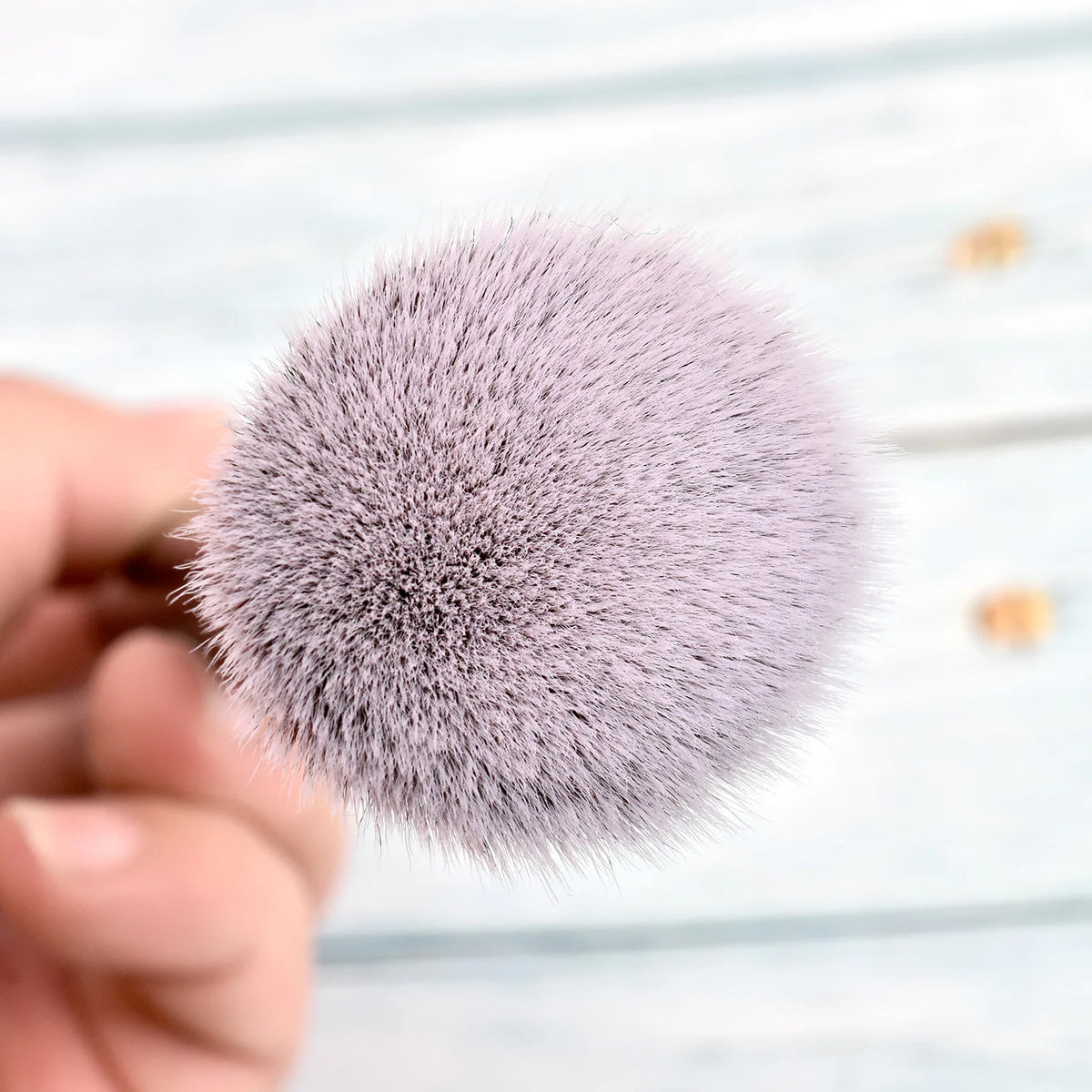 A close-up of a fluffy, soft lavender makeup brush being held by a hand against a light wooden background.
