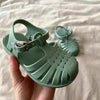 A hand holds a pair of mint green kids' Roman Gladiator jelly sandals with non-slip soles.