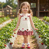 A young girl wearing a white pinafore dress with strawberry and bunny prints stands among green plants.