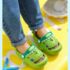 A pair of bright green kids shark clog sandals with anti-slip soles resting on a yellow surface.