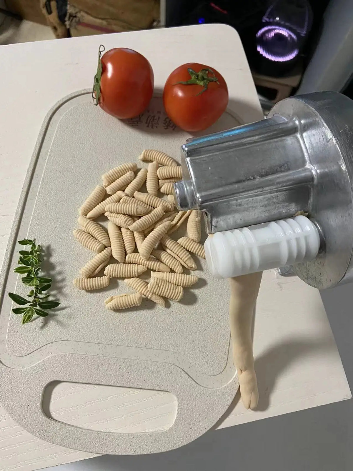 A silver pasta maker machine is shown next to fresh tomatoes and a pile of dry cavatelli pasta.