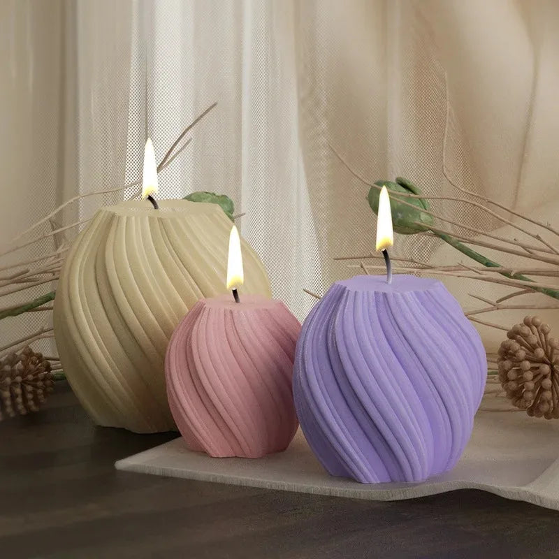 A purple pillar candle with a flame sits on a white surface next to a large beige candle and dried branches.