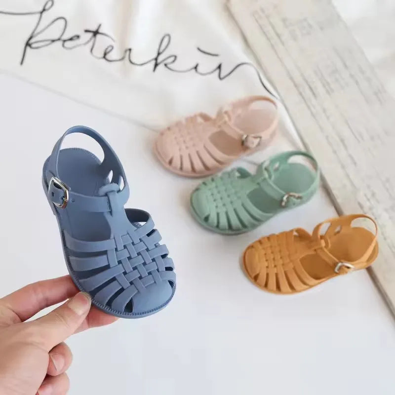 A hand holds a pair of kids' Roman gladiator jelly sandals in various pastel colors.