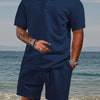 A man wearing a navy blue jacquard short-sleeve shirt and matching drawstring shorts set standing near the water.