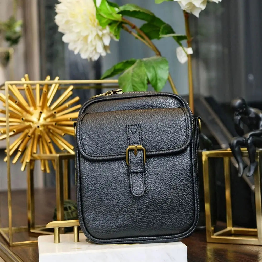 Black vegan leather messenger bag with gold hardware displayed on a marble surface.