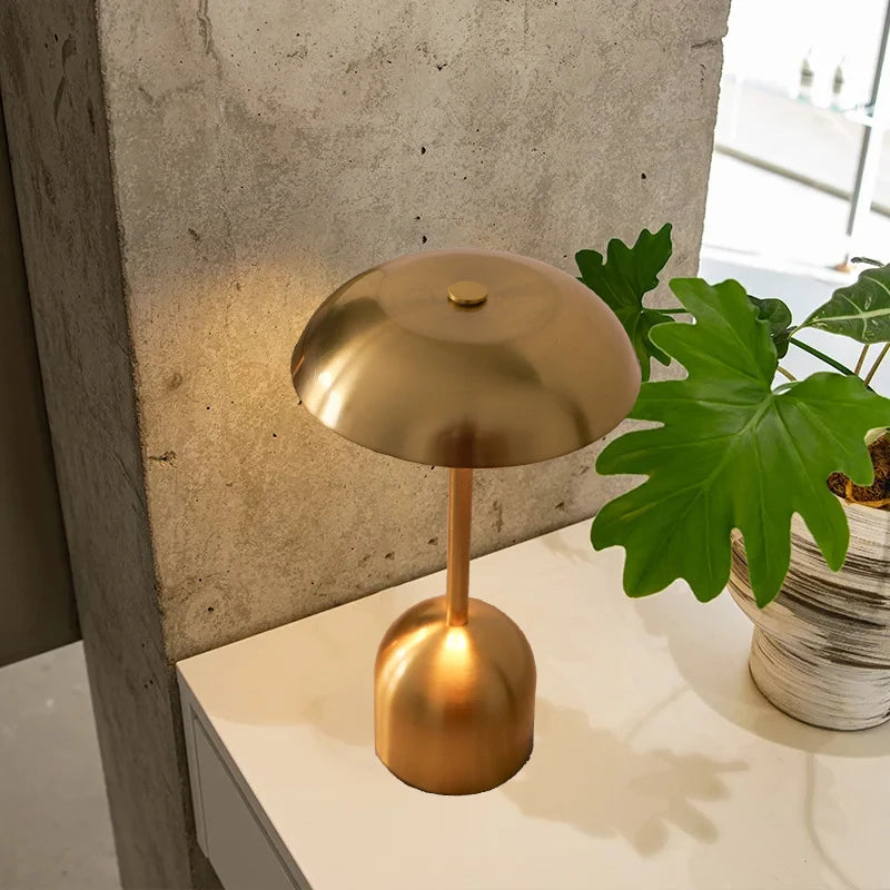 A minimalist gold-toned touch-control rechargeable LED table lamp with a dome shade sits on a white surface.