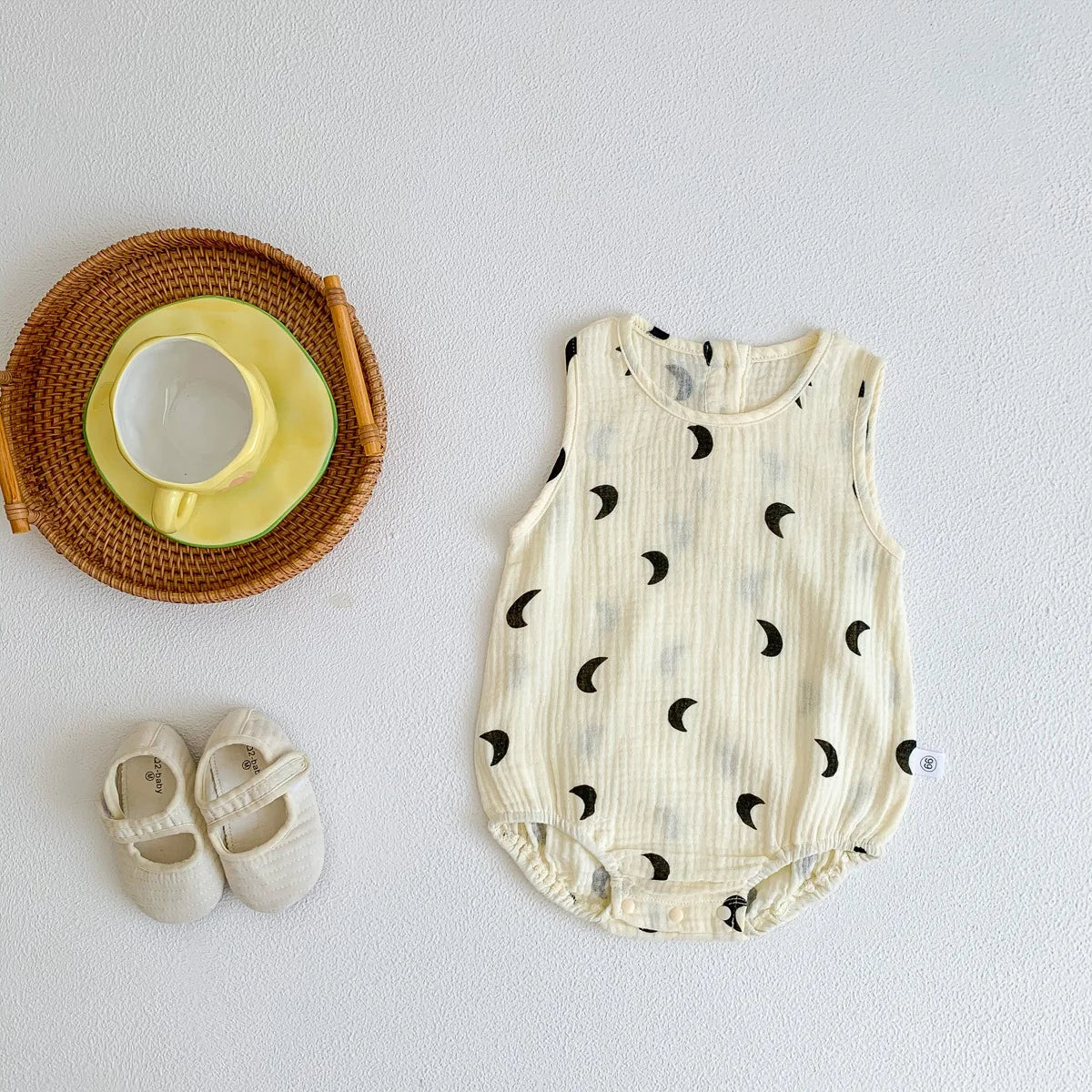 A white baby cotton sleeveless jumpsuit bodysuit set with black crescent moon patterns, next to a yellow bowl and wooden spoo