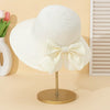 A white straw sun hat with a large bow is displayed on a gold stand against a light background.