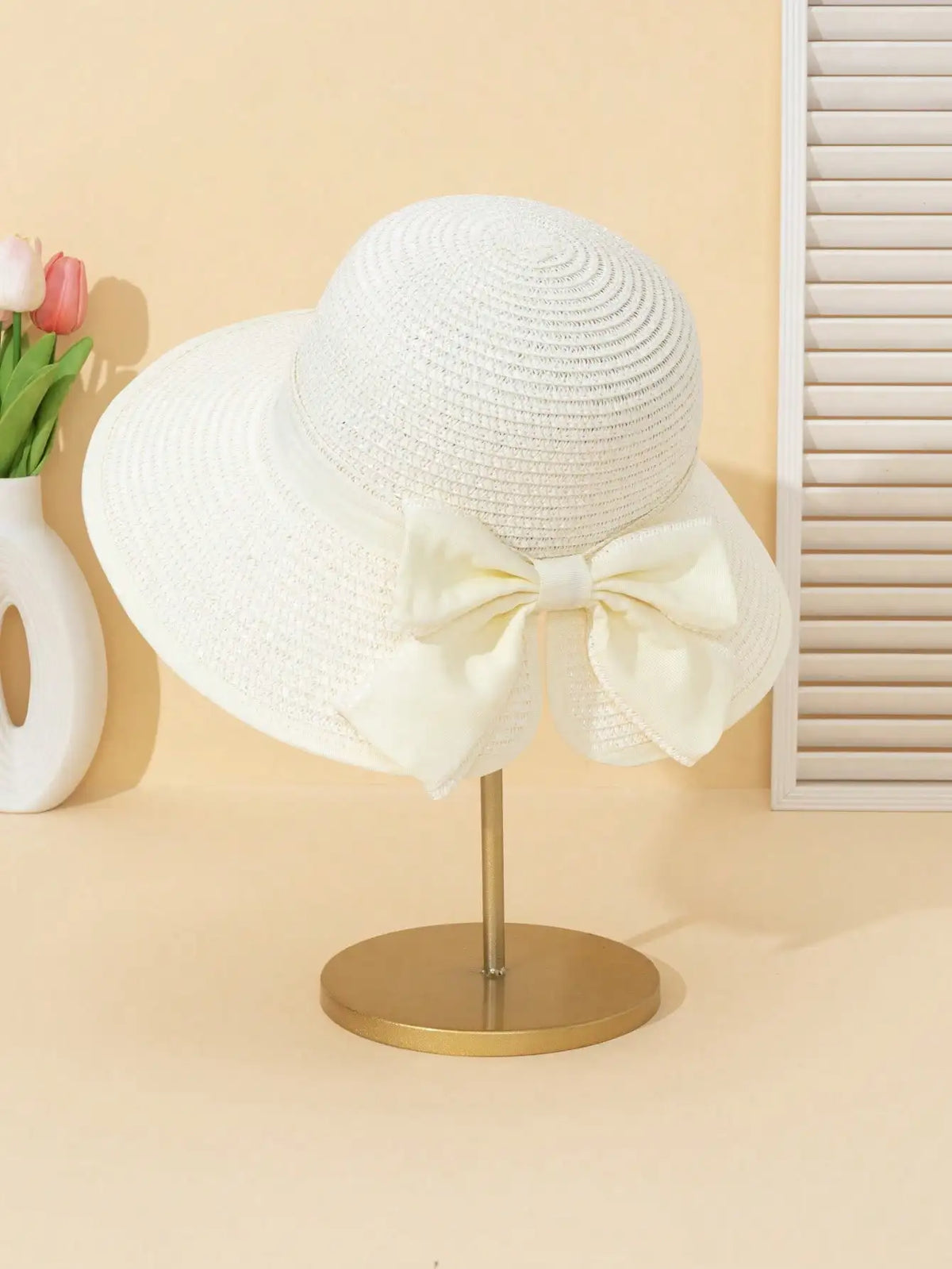 A white straw sun hat with a large bow is displayed on a gold stand against a light background.