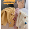 A display of yellow, pink, and cream knitted sweaters with pom-pom trims next to a wooden bear shape.