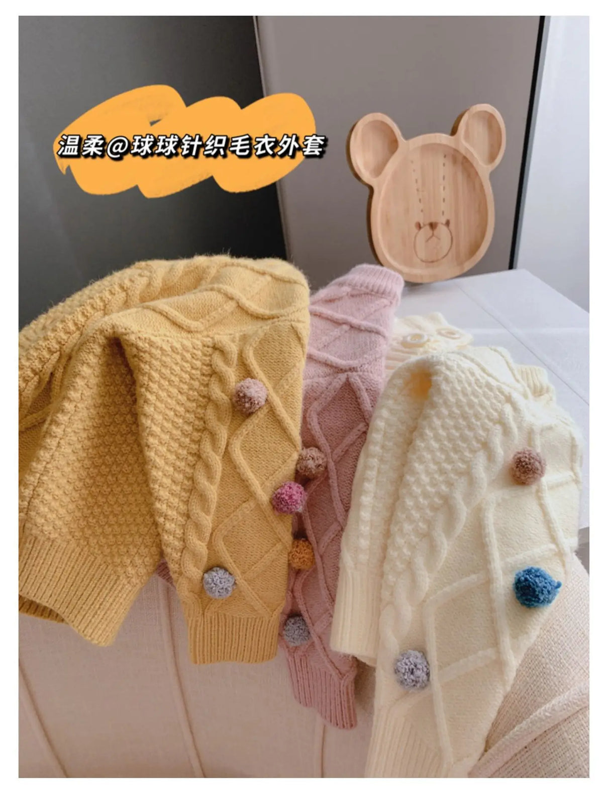 A display of yellow, pink, and cream knitted sweaters with pom-pom trims next to a wooden bear shape.