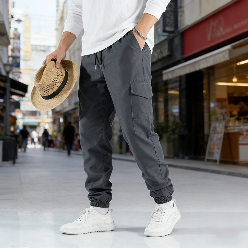 A man wearing gray casual cargo pants and white sneakers is walking on a city sidewalk.
