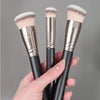 A hand holds three precision makeup brushes with fluffy pink and white bristles and silver ferrules.