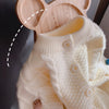 A close-up of a cream knitted cardigan sweater with a high neck and pom-pom trim, shown next to a wooden hanger.