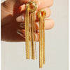 A pair of gold geometric tassel drop earrings held in a person's hand.