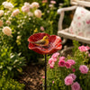 A small yellow bird sits on a red metal flower, surrounded by pink and white roses in a garden setting.