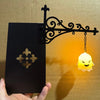 A black decorative lantern featuring an ornate wrought iron bracket and a glowing yellow light bulb.