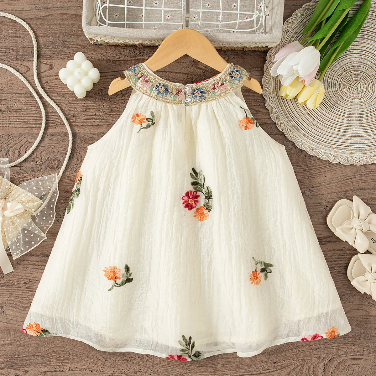 A cream-colored floral embroidered sleeveless dress for baby girls displayed on a wooden hanger.