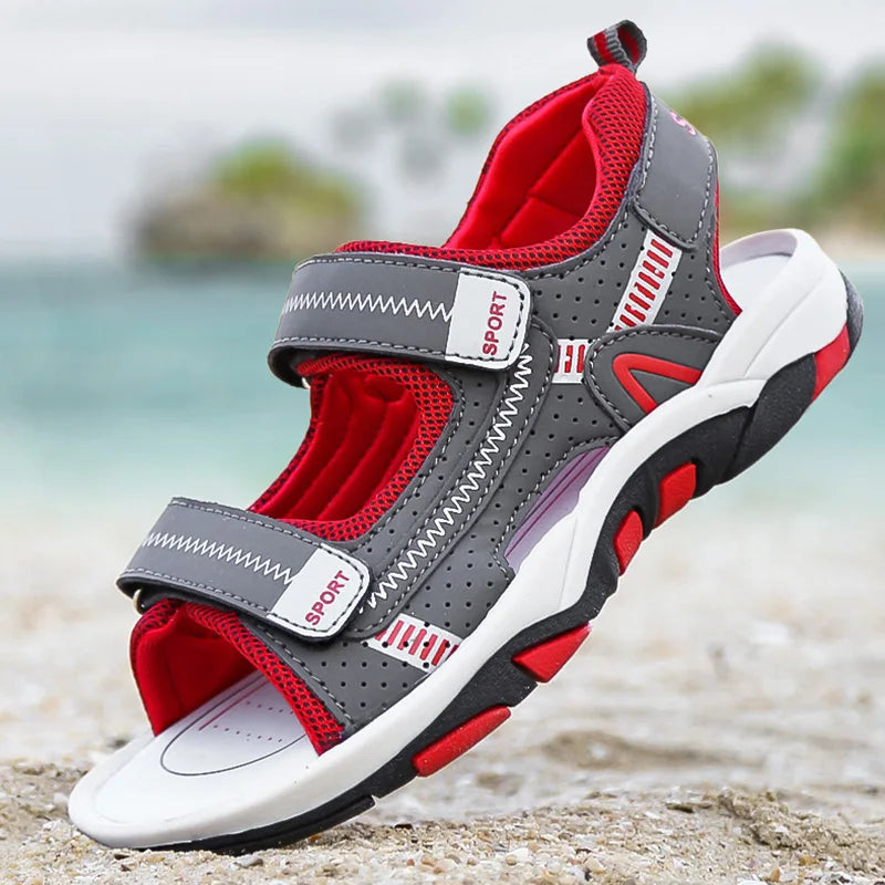 A pair of gray and red breathable summer sport sandals with adjustable straps resting on a sandy beach.