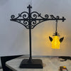 A black wrought iron shelf lamp featuring a decorative cross and a hanging glass lantern with a warm yellow glow.