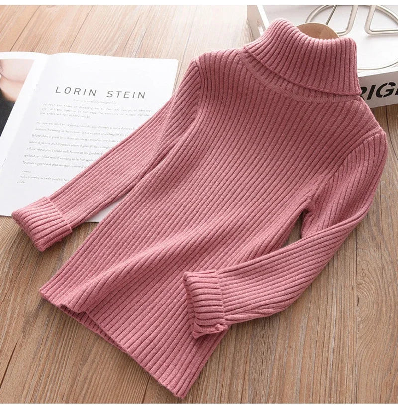 A pink ribbed knit turtleneck sweater laid flat on a wooden surface next to a book.