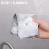 A hand holds a white foaming cleaning cloth with silver accents, demonstrating rich foaming action.