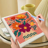 Hands are using a needle to embroider a colorful flower pattern on a canvas within a framed kit.