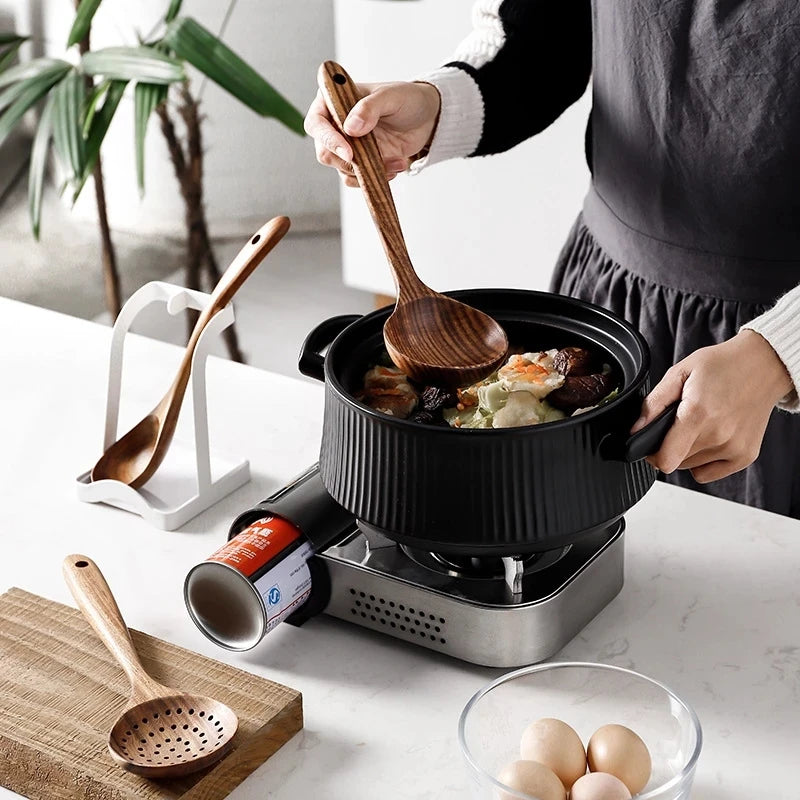A person is using a natural teak wood cooking utensil, such as a spoon or spatula, in a black non-stick pan on an electric co