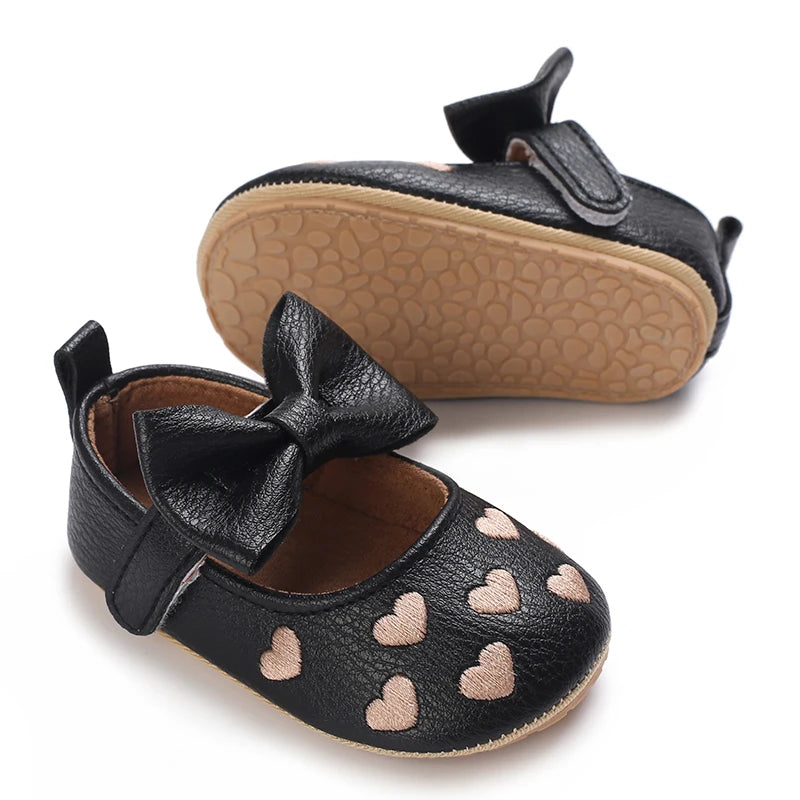 A pair of black baby first walker shoes with a bow and heart pattern on the toe