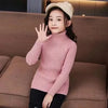 A young girl wearing a pink knit turtleneck sweater and a beige knit hat sitting on a brown sofa.