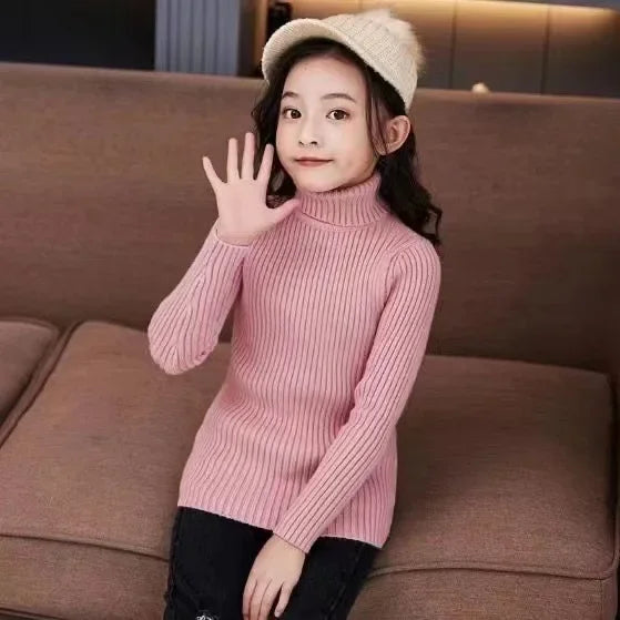 A young girl wearing a pink knit turtleneck sweater and a beige knit hat sitting on a brown sofa.