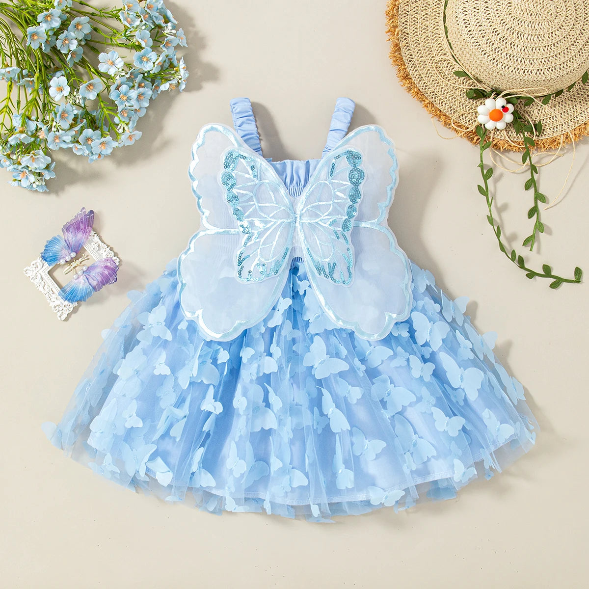 A light blue tulle butterfly dress for a baby girl displayed on a beige background with flowers and a straw hat.