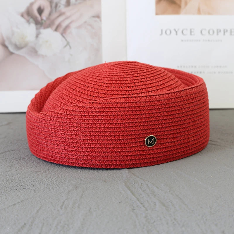 A close-up of a red knitted straw pillbox sun hat with a wide brim resting on a gray surface.