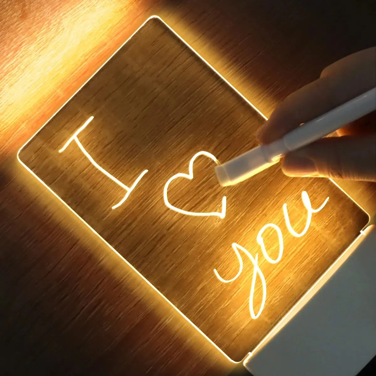 A hand writes I love you on a glowing acrylic message board illuminated by a desk lamp.