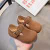 A pair of brown suede moccasin shoes with leather straps and hook-and-loop closure being held in a hand.