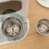 A close-up of a double-wall insulated stainless steel espresso cup and a small metal cup on a wire rack.
