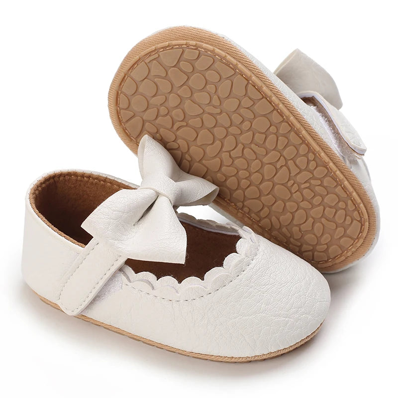 A pair of white soft non-slip baby first walker shoes with light brown textured soles and bow details.