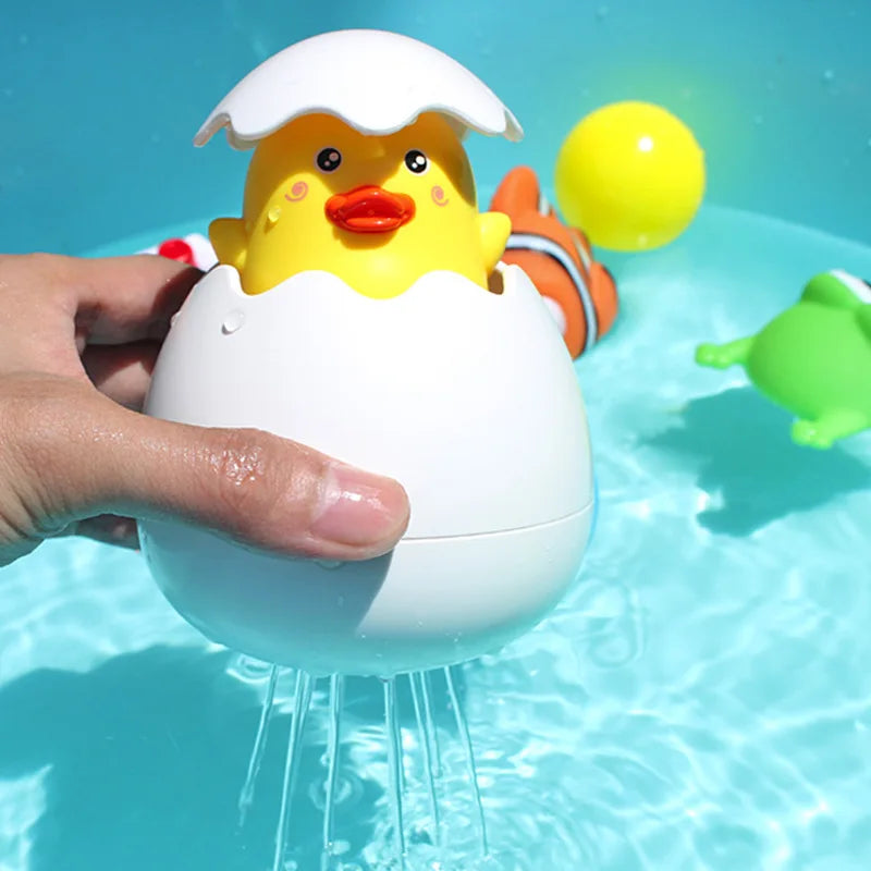 A hand holds a yellow duck bath toy with a white egg shape, spraying water into a blue pool.
