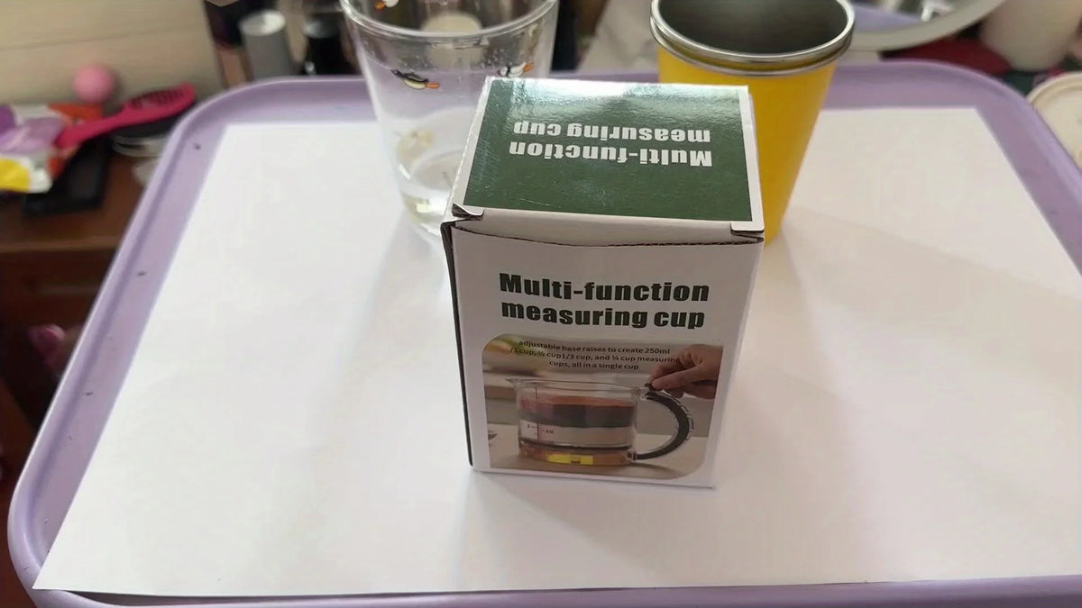 A box of a multi-function measuring cup is displayed on a white surface next to a glass and a yellow container.