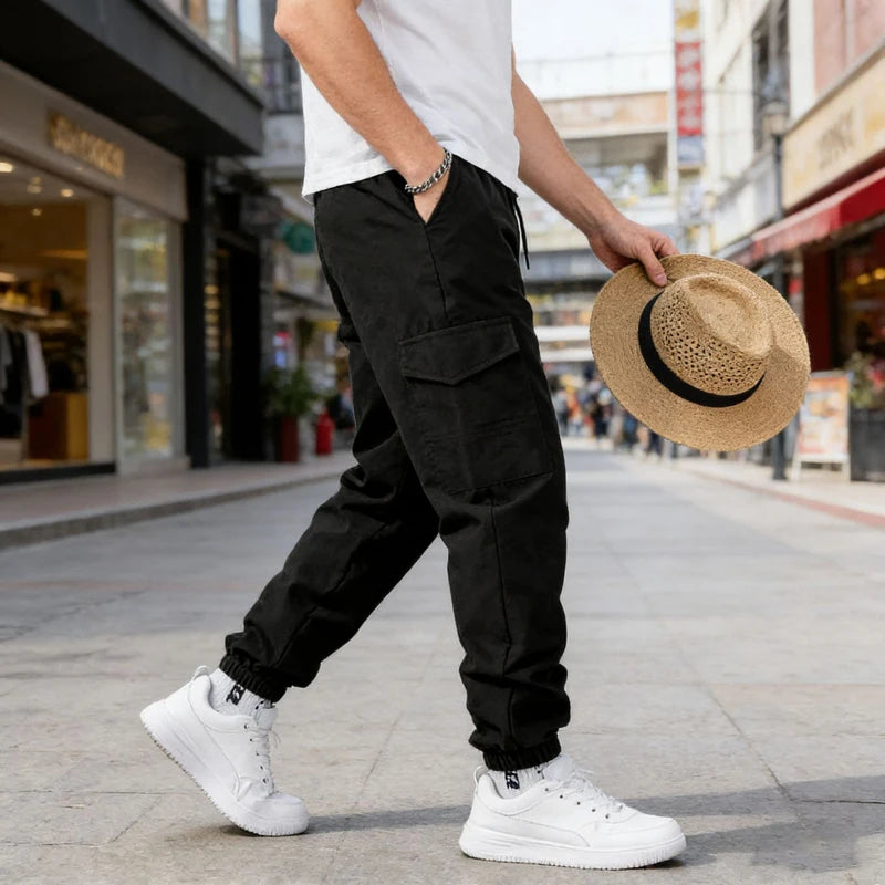 A man wearing black casual cargo pants and a white shirt walks on a city sidewalk while holding a straw hat.