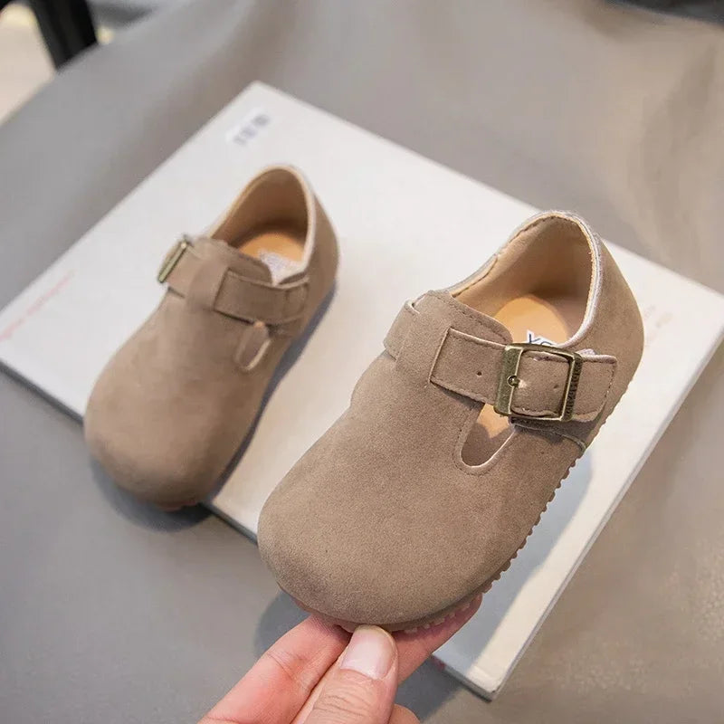 A pair of tan suede kids' moccasin shoes with a hook-and-loop closure resting on a white surface.