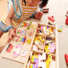 A child is interacting with a wooden puzzle set featuring bear dress-up elements and various small wooden toys.