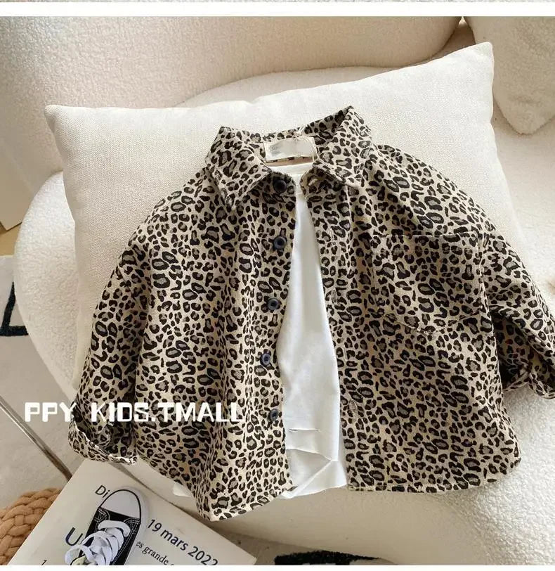 A beige leopard print denim jacket is displayed on a white cushion with a book and sneakers in the background.