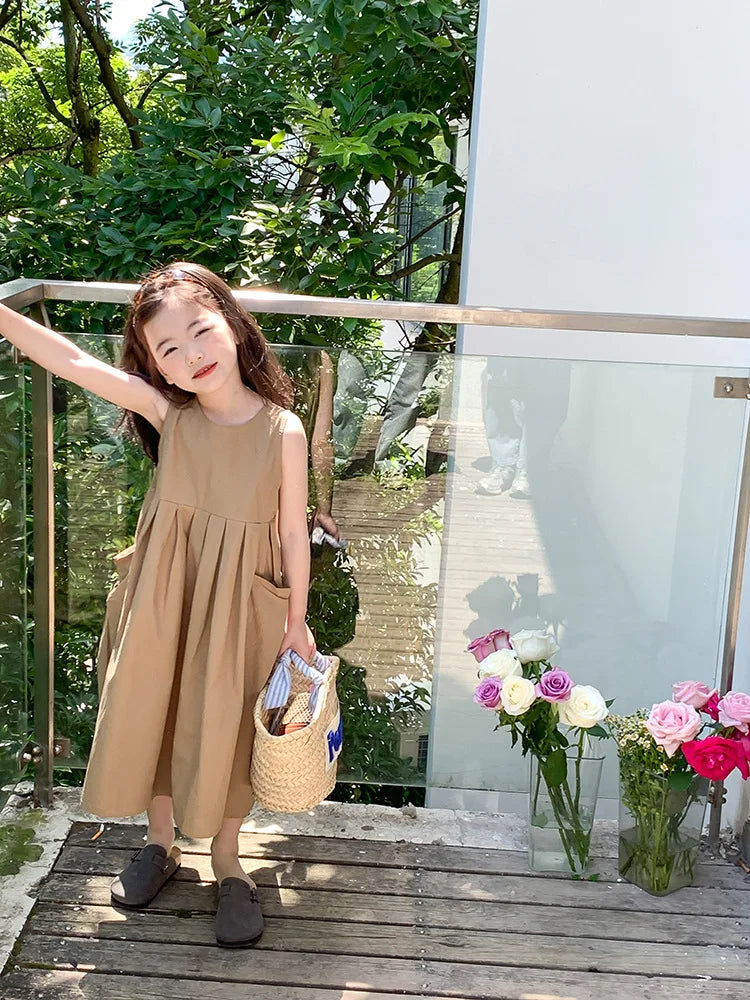 A young girl wearing a beige cotton sleeveless sundress standing outdoors near a glass railing.