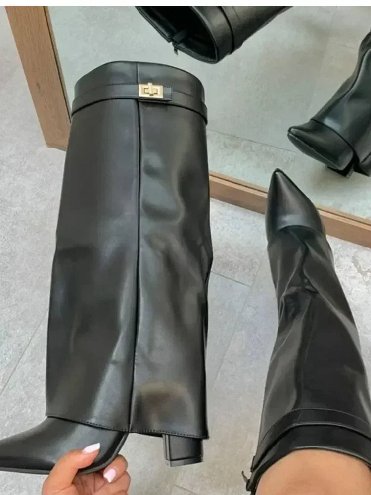 A black knee-high chunky heel boot with a metal buckle is shown on a person's foot.