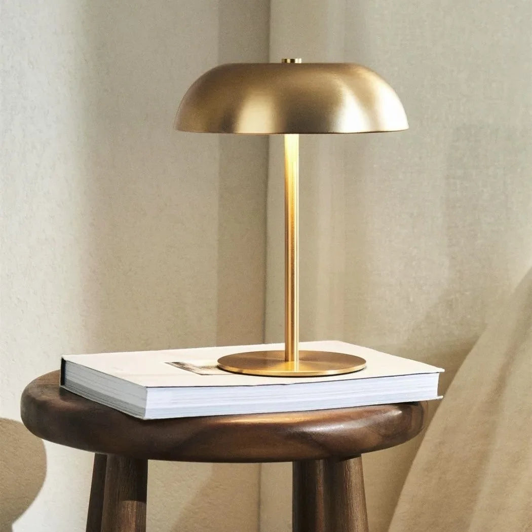A modern brass portable touch table lamp with a dome shade rests on a stack of white books.