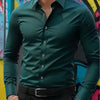 A man wearing a dark green long-sleeve formal shirt and black pants, posing in front of colorful graffiti.