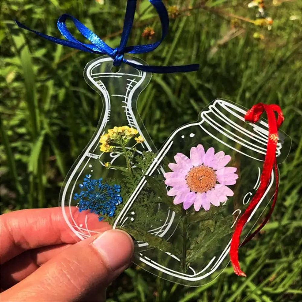 A hand holds a clear glass bookmark featuring dried flowers and a red ribbon against a green outdoor background.